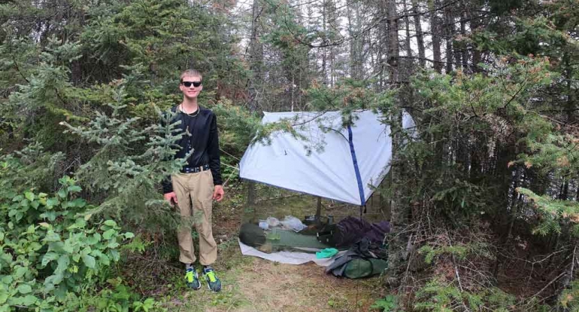 a student stands beside their tarp shelter in a wooded area on an outward bound trip
